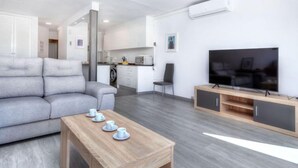Una sala moderna con un sofá gris, una mesa de centro de madera con tazas, un televisor sobre un mueble de madera y una cocina al fondo.
