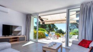 Una sala de estar moderna con un televisor de pantalla plana, sofás grises, una mesa de centro de madera y un puerta corrediza de vidrio que da a un patio exterior con sillas y una mesa.