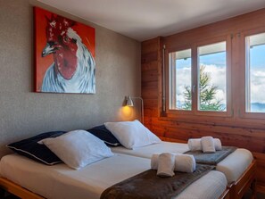 A room with two beds, a painting of a rooster, a window with a view of trees, and a lamp. - Chalet Aquarius by Interhome (Nendaz)