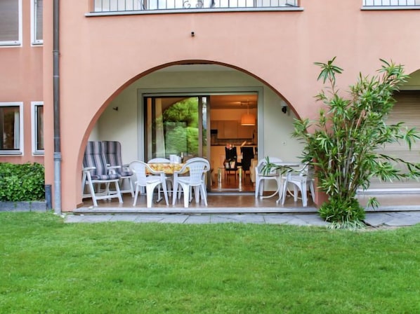 A building with a covered outdoor seating area featuring white chairs and a table, surrounded by greenery.