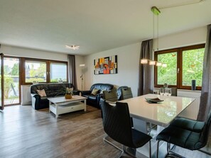 A modern living room with a sofa, coffee table, and dining table. There are large windows with curtains, a ceiling light, and a wall-mounted artwork. - Vacation home Quelle by Interhome (Dittishausen)