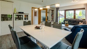 Ein moderner Essbereich mit einem weißen Tisch, schwarzen Stühlen und einem blauen Sofa. Ein Bücherregal und ein Fenster mit Blick auf Grün sind ebenfalls vorhanden.