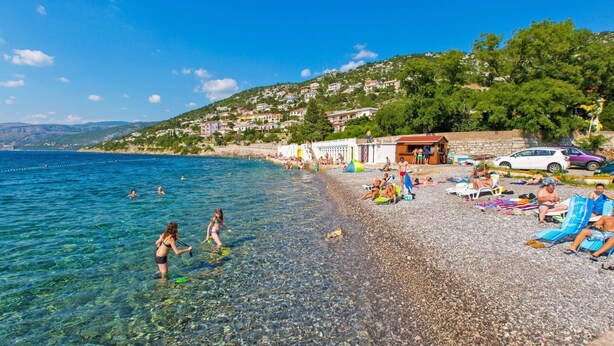 Une plage de galets avec une eau claire, des gens qui se baignent et des chaises de plage.