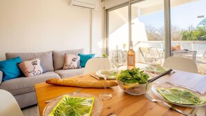 A modern living room with a wooden dining table set for a meal, a grey sofa with patterned cushions, and large windows offering a view of the outdoors. - du Parc I by Interhome (La Grande Motte)