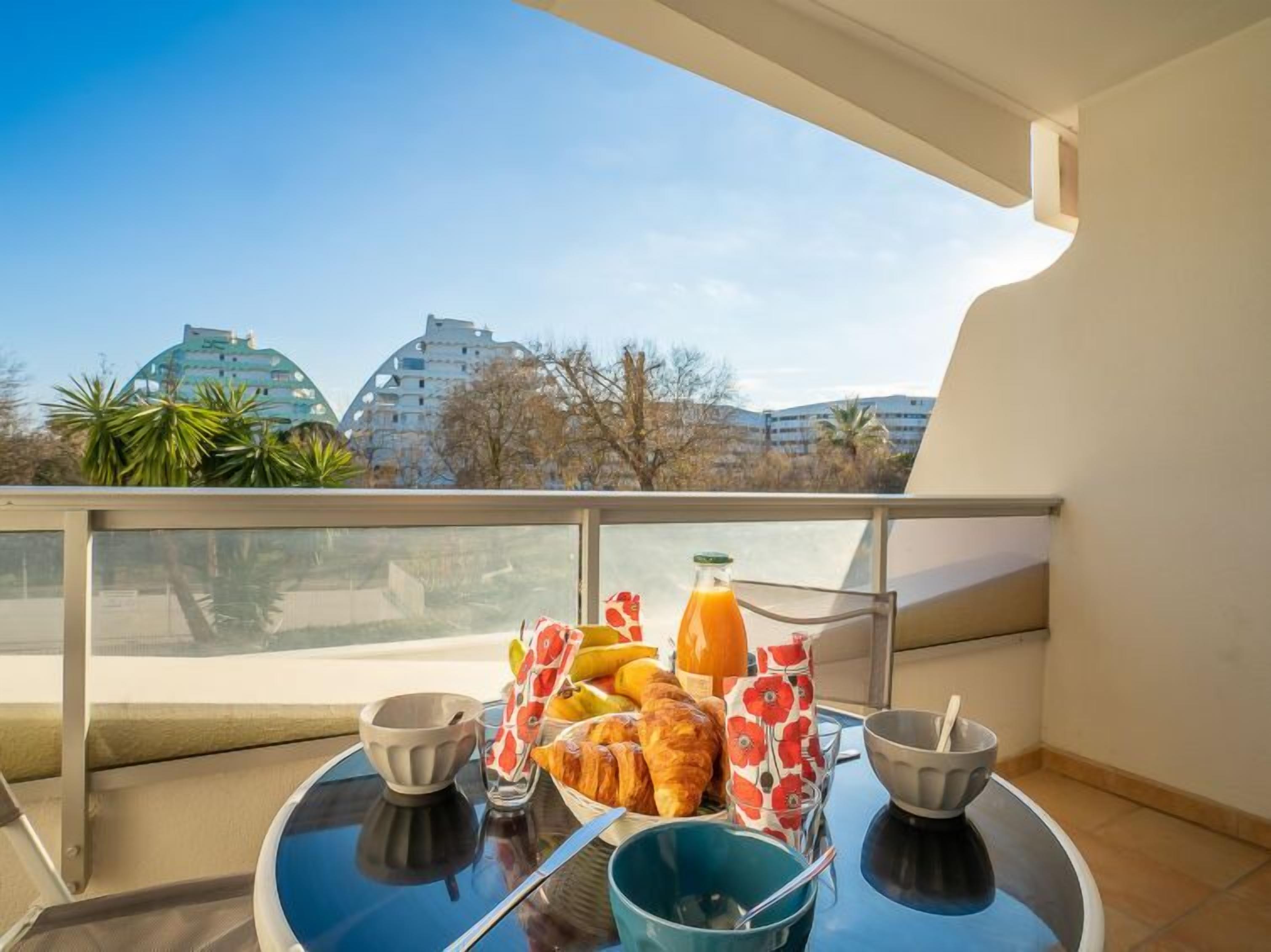 Ein Balkon mit einem für das Frühstück gedeckten Tisch, darunter Croissants, eine Flasche Orangensaft und ein Obstkorb.