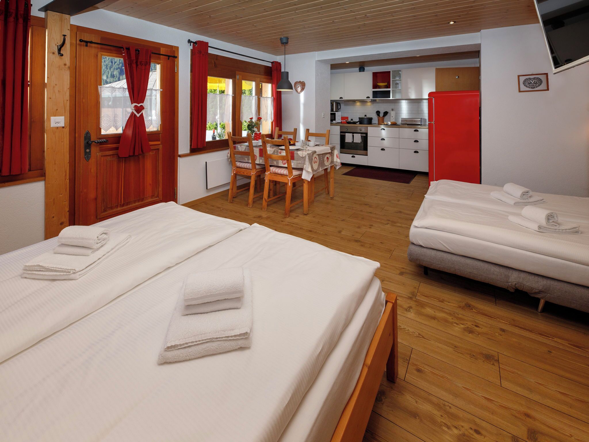 A hotel room with two beds, a dining table, chairs, a kitchen area, and a red wall.