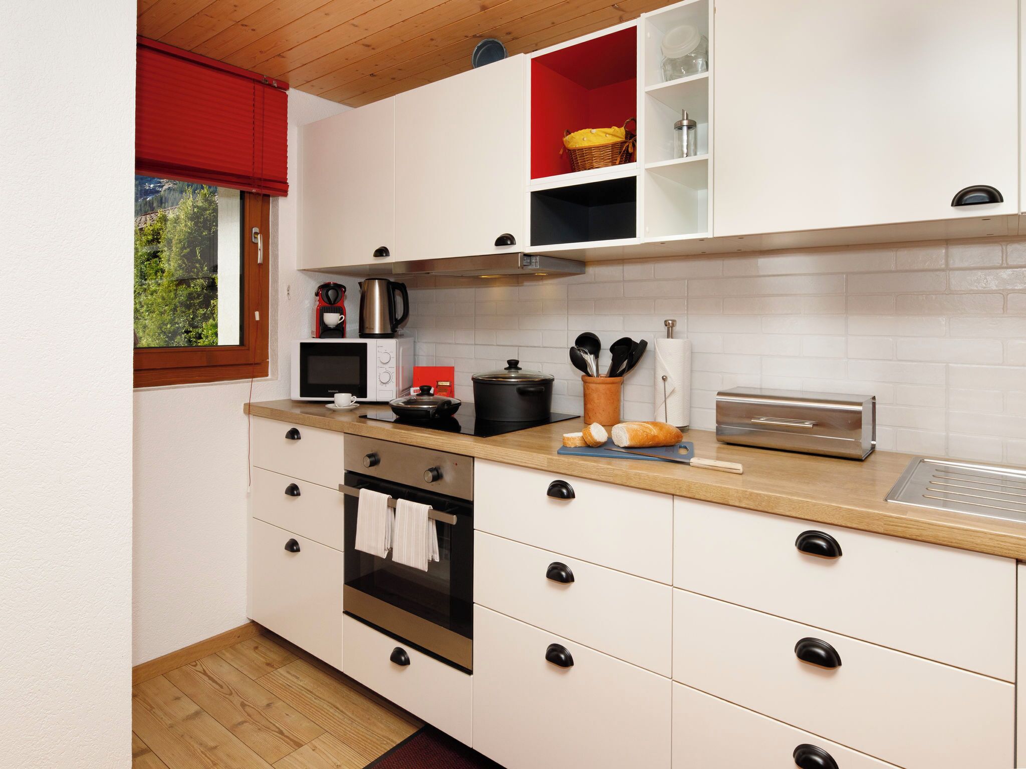A modern kitchen with white cabinets, a wooden countertop, a stainless steel oven, and a red window blind.