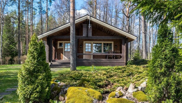 Una cabaña de madera con un porche, rodeada de árboles y un área verde con rocas cubiertas de musgo.