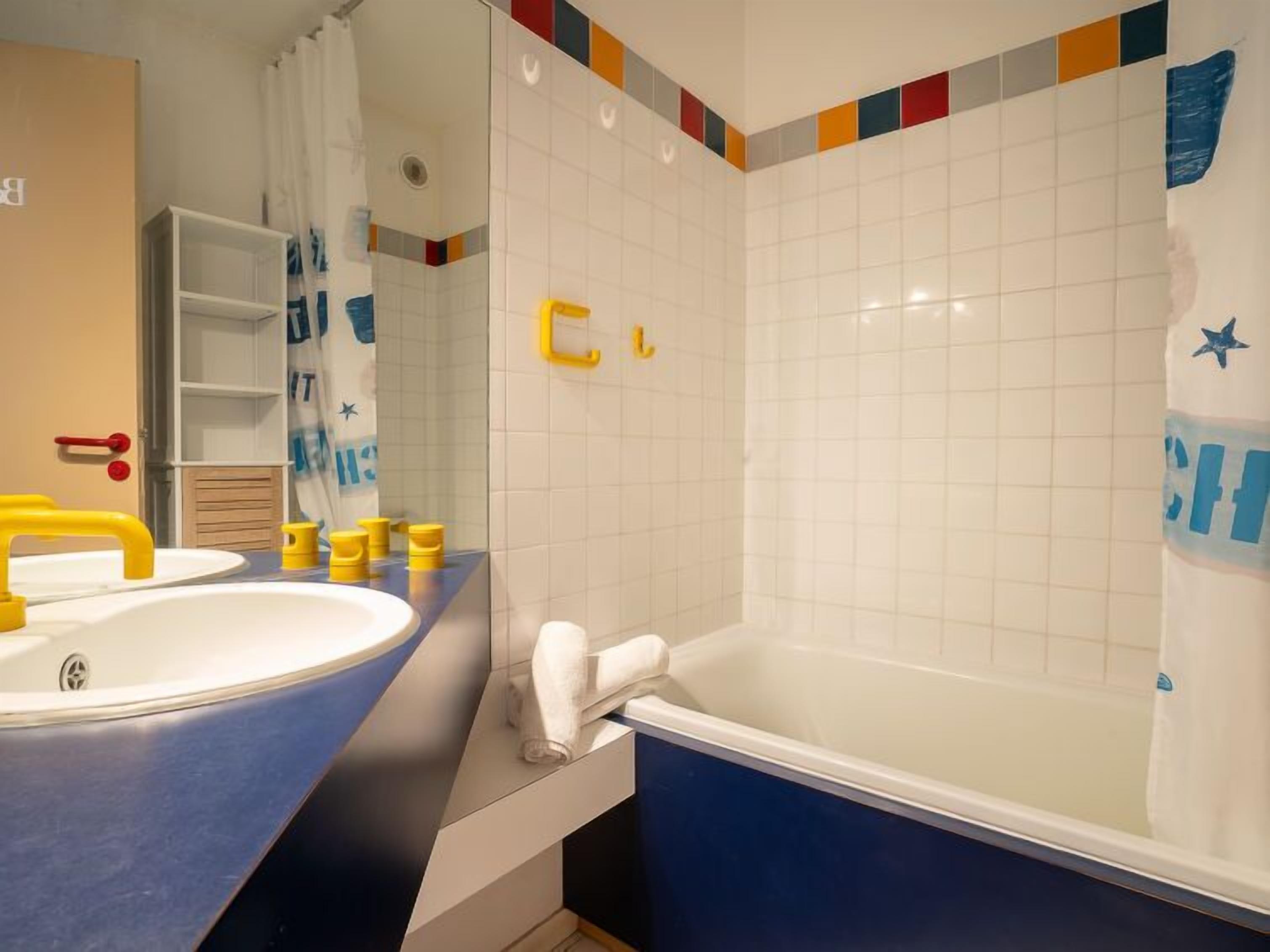 A bathroom with a bathtub, a sink, and a mirror. There are colorful tiles on the walls and a shelf with toiletries.