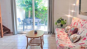 Ein Wohnzimmer mit einer Couch, einem kleinen Tisch und einem Sessel. Eine Schiebetür führt zu einem Außenbereich mit Tisch und Stühlen.