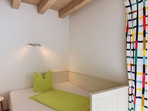 Una habitación pequeña con cama individual, cabecera blanca, almohada verde, colchón blanco y cortina a rayas de colores.