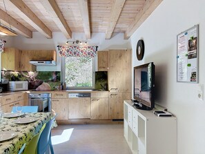 Cocina moderna con armarios de madera, un comedor con mesa para dos personas y un amplio ventanal con vistas al verdor.