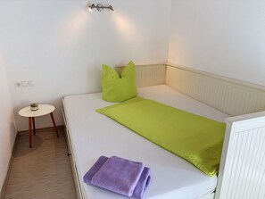 Une chambre avec un lit simple, une tête de lit blanche, une petite table ronde avec un récipient en verre et un coussin vert.