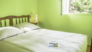 Uma cama com cabeceira de madeira, uma mesa de cabeceira com abajur e uma janela com vista para o verde lá fora.