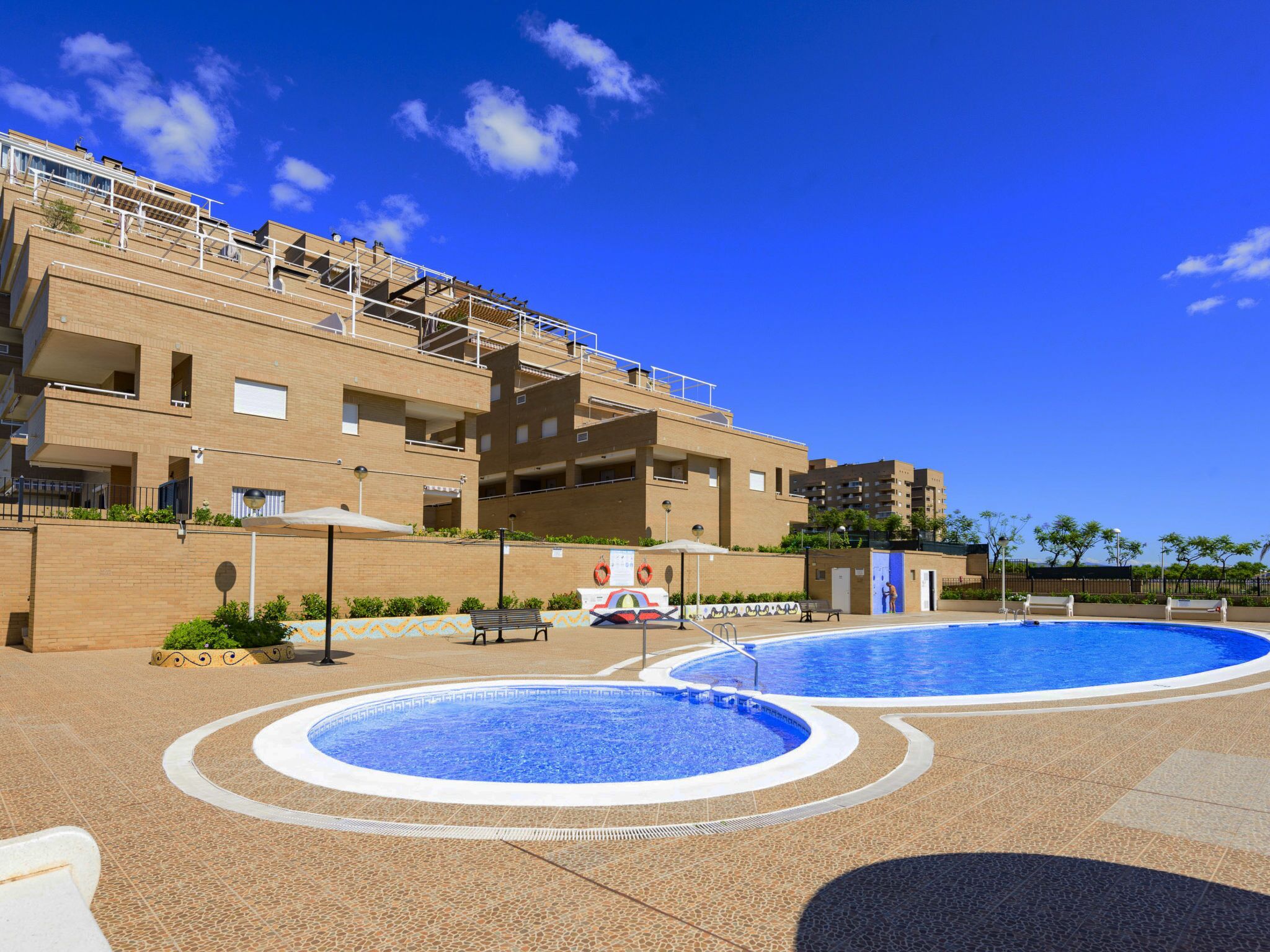 A pool area with two circular pools, a brick building with balconies, and a clear blue sky.
