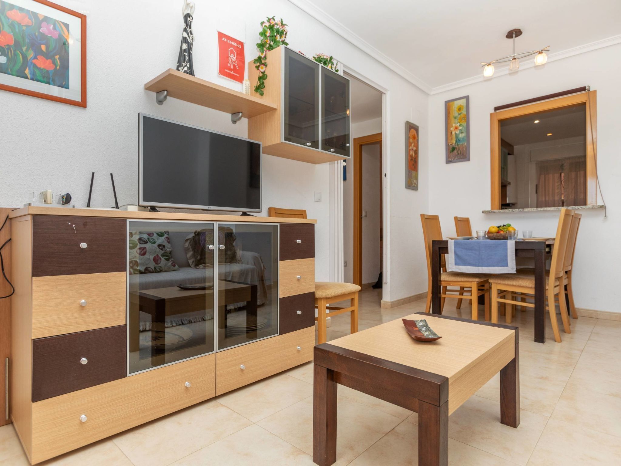 A modern living room with a TV, wooden cabinet, glass doors, a dining table with chairs, and a kitchen area visible through an open door.