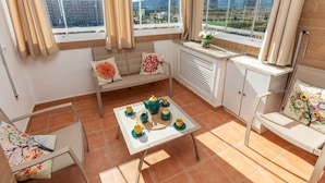 Ein helles Zimmer mit einem großen Fenster, einem Sofa, einem Tisch mit Tassen und einem Teekessel sowie einem Sessel mit einem Blumenkissen.