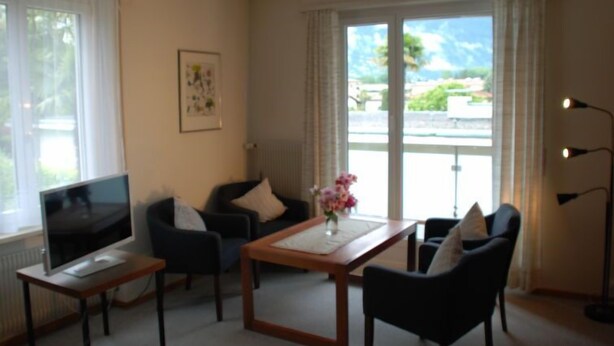 A living room with a TV, two chairs, a table, and a window with a view of greenery and buildings. - Residenza Corallo by Interhome (Ascona)