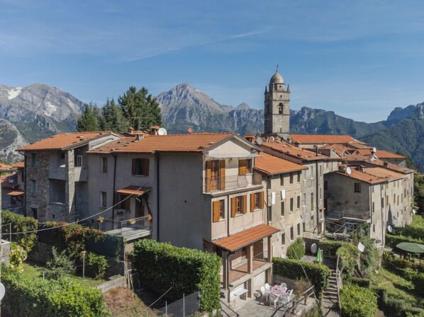 A traditional stone building with a bell tower, surrounded by lush greenery and mountains.