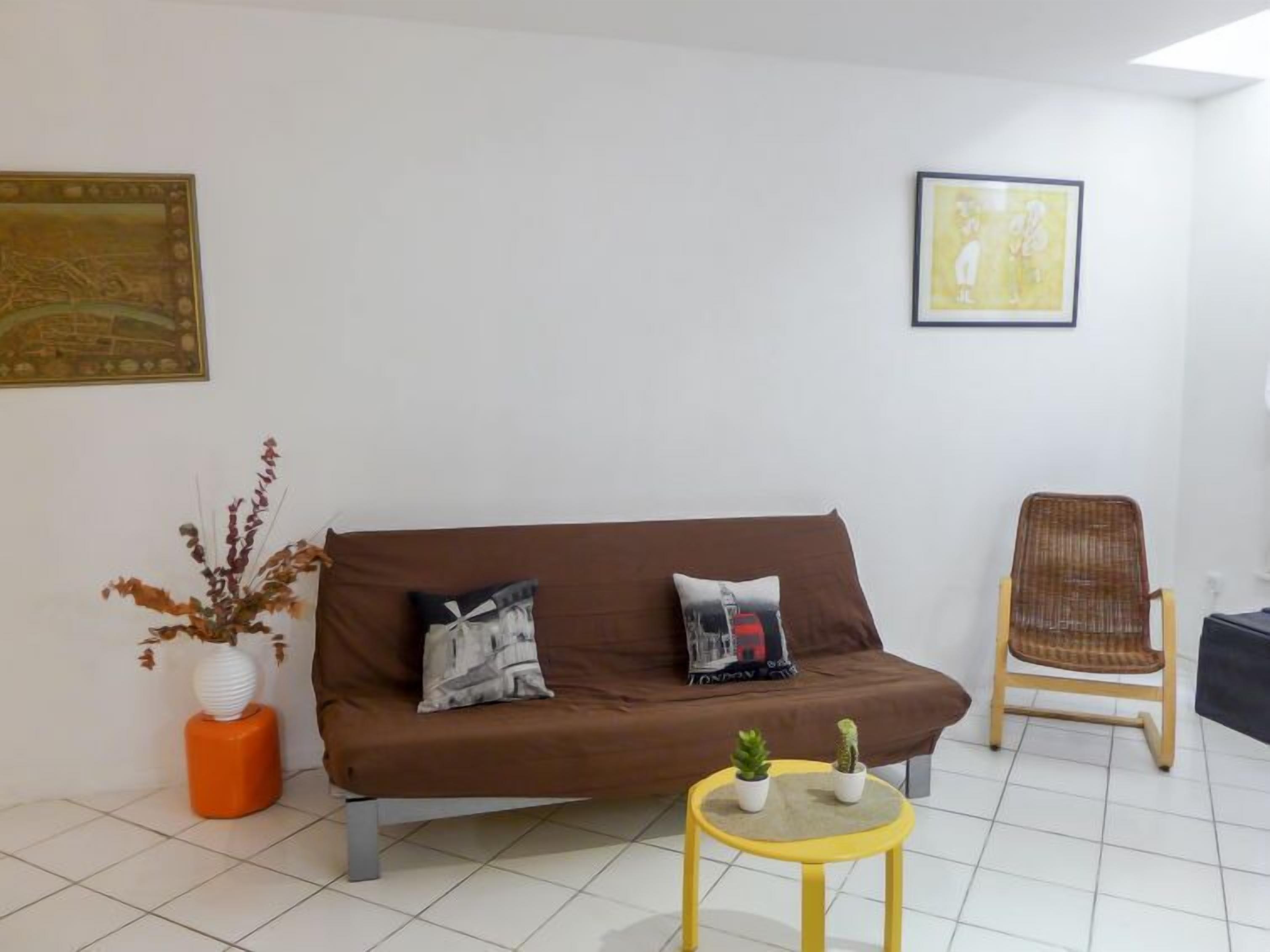A living room with a brown sofa, two cushions, a small round yellow table with two potted plants, a wicker chair, and two framed artworks on the walls.