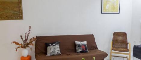 A living room with a brown sofa, two cushions, a small round yellow table with two potted plants, a wicker chair, and two framed artworks on the walls.