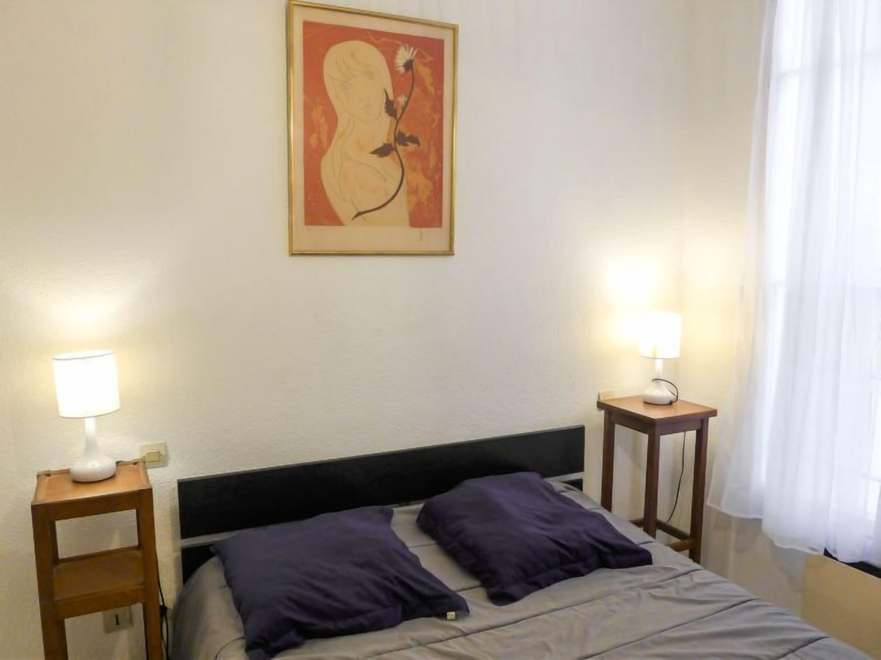 Ein Hotelzimmer mit einem Bett, zwei Nachttischen mit Lampen, einem gerahmten Kunstwerk an der Wand und einem Fenster mit Vorhängen.