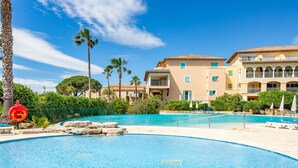 A swimming pool with a clear blue water, surrounded by a sandy beach and palm trees. In the background, there is a multi-story building with balconies and a tiled roof. - Les Rivages des Issambres by Interhome (Les Issambres)