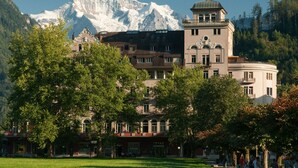 Un edificio storico con una torre, circondato da alberi rigogliosi e con montagne sullo sfondo.