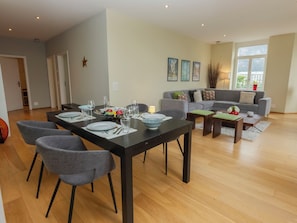 A modern living room with a dining area, grey sofas, a grey dining table set for four, and a large window with a view.