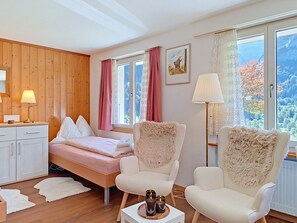 Een slaapkamer met een bed, twee stoelen, een nachtkastje, een lamp en een raam met gordijnen.