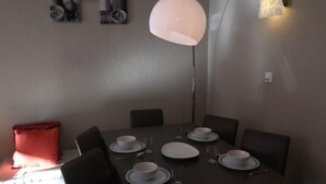 Ein Essbereich mit Tisch für vier Personen, gedeckt mit Tellern, Schüsseln und Besteck. Zwei Wandleuchten und ein rotes Kissen liegen auf dem Boden.