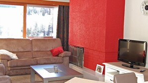 Ein Wohnzimmer mit einem braunen Sofa, einer roten Akzentwand, einem Kamin, einem Fernseher und einem Fenster mit Blick auf Bäume.