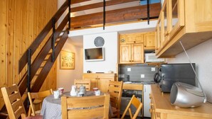 A compact kitchen with wooden cabinets, a dining area with a table and chairs, a clock, and a microwave. - Asters 1208 Batiment B4 by Interhome (Les Menuires)