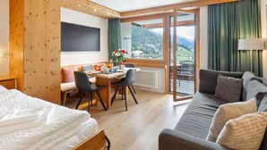 Habitación de hotel con cama, sofá, mesa de comedor con sillas, kitchenette y balcón con vistas a la montaña.