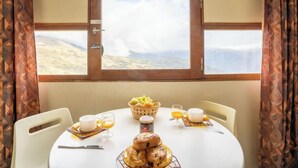 A dining table with breakfast items, a view of a mountainous landscape through a window, and patterned curtains. - Vostok Zodiaque by Interhome (Le Corbier)
