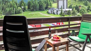 Un balcón con una mesa, sillas y una botella de vino. Se ve una vista de la montaña con un edificio al fondo.