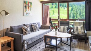 Una zona de estar acogedora con un sofá gris, dos sillones, una mesita de centro y una lámpara de pie. Un cuadro cuelga de la pared, y una ventana ofrece vistas a una zona boscosa.