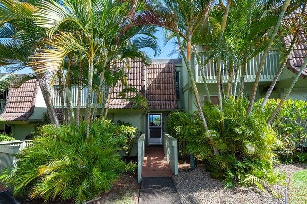 Un immeuble à plusieurs étages, doté d’une façade verdoyante et d’un toit brun, entouré d’une végétation tropicale luxuriante.