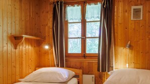 A wooden cabin room with two beds, a window with curtains, a wall-mounted lamp, and a small heater. - Coquelicot by Interhome (Zinal)
