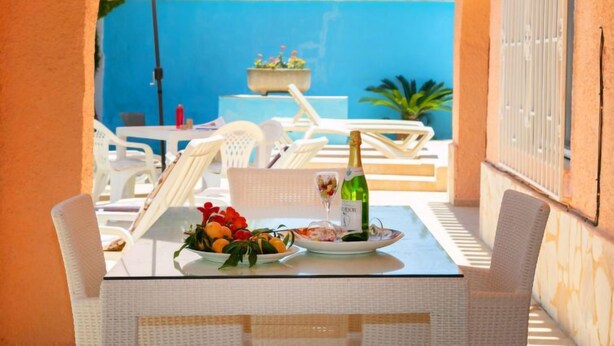 A table set with a bottle of champagne, a glass, and a plate of fruit, surrounded by white chairs and an umbrella, with a blue wall and a plant in the background. - Vacation home Sandra by Interhome (Albir)