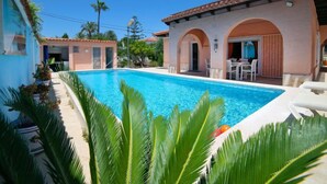 A swimming pool with a clear blue water, surrounded by palm trees and a building with a tiled roof and arches. - Vacation home Sandra by Interhome (Albir)