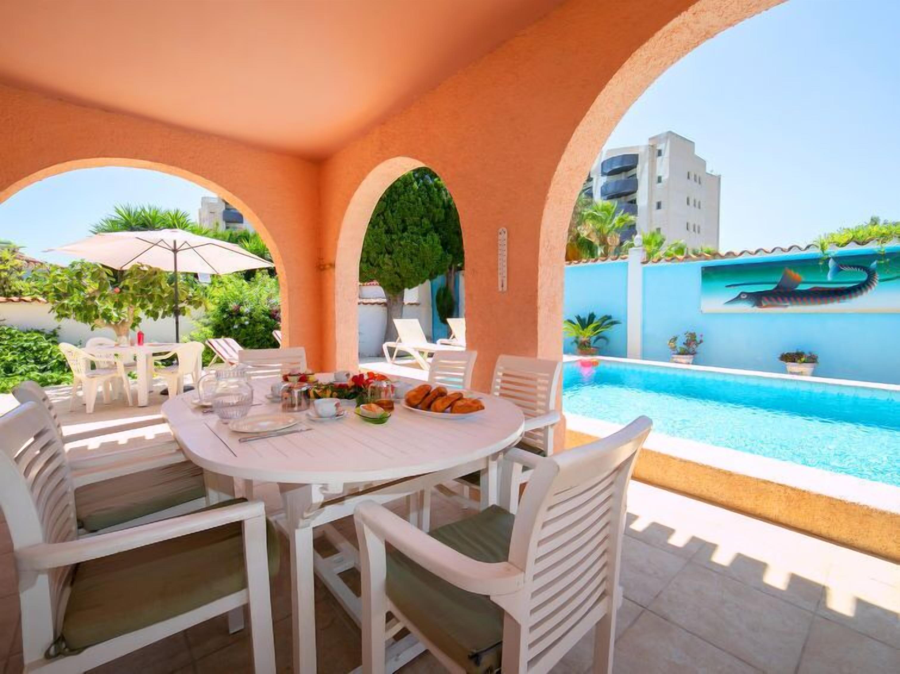 Un espace repas aménagé au bord de la piscine, avec des meubles d’extérieur blancs, une table dressée avec des assiettes et des verres, et une vue sur une piscine et des bâtiments en arrière-plan.