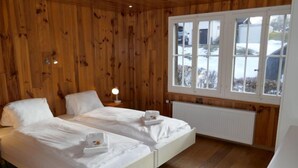 Une chambre à coucher aux murs lambrissés, avec deux lits, une table de chevet dotée d’une lampe et une fenêtre donnant sur l’extérieur.