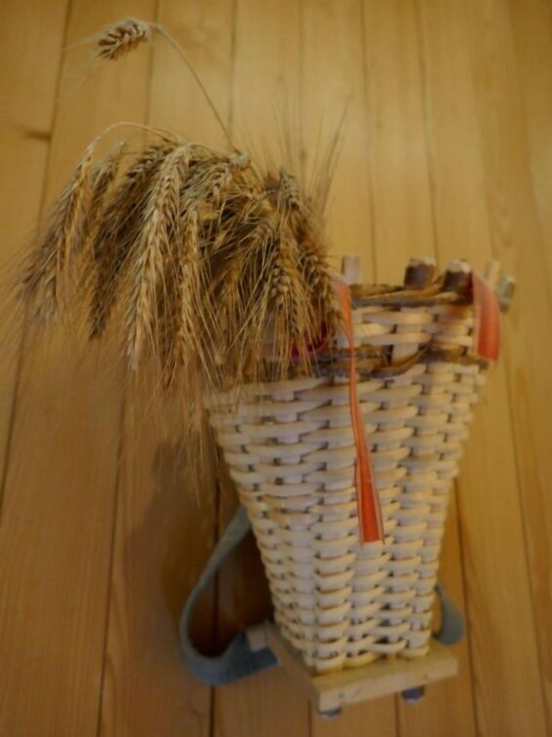 A woven basket with a handle, filled with dried wheat stalks, placed on a wooden floor.