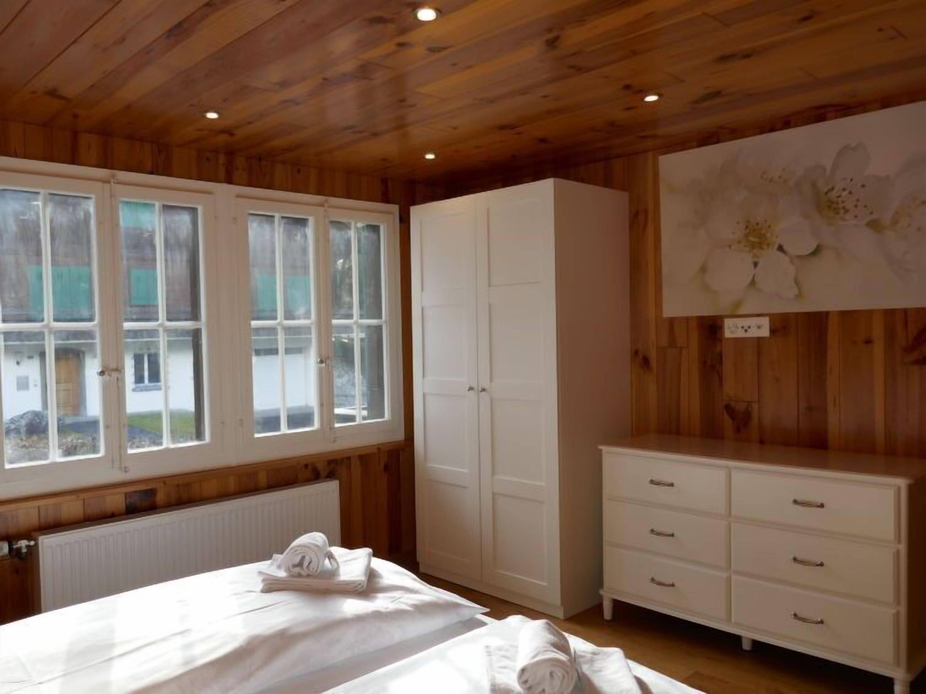Ein Schlafzimmer mit Holzwänden, einem Bett mit weißen Leinen, einer weißen Kommode und einem großen Fenster mit Außenblick.