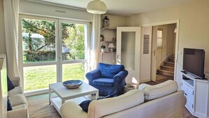 Een woonkamer met een bank, een blauwe fauteuil, een salontafel en een televisie. Er is een groot raam met uitzicht op groen.