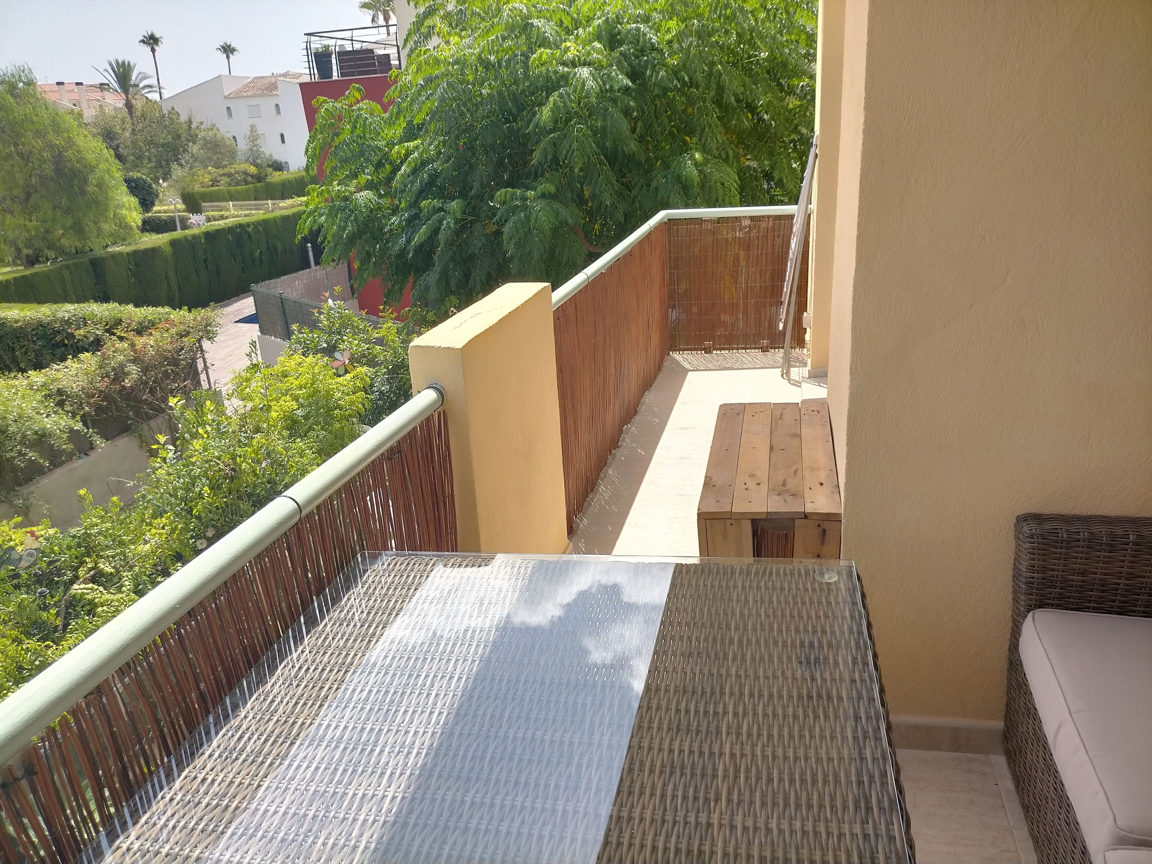 A balcony with a wicker sofa, a wooden table, and a view of a garden with hedges and trees.