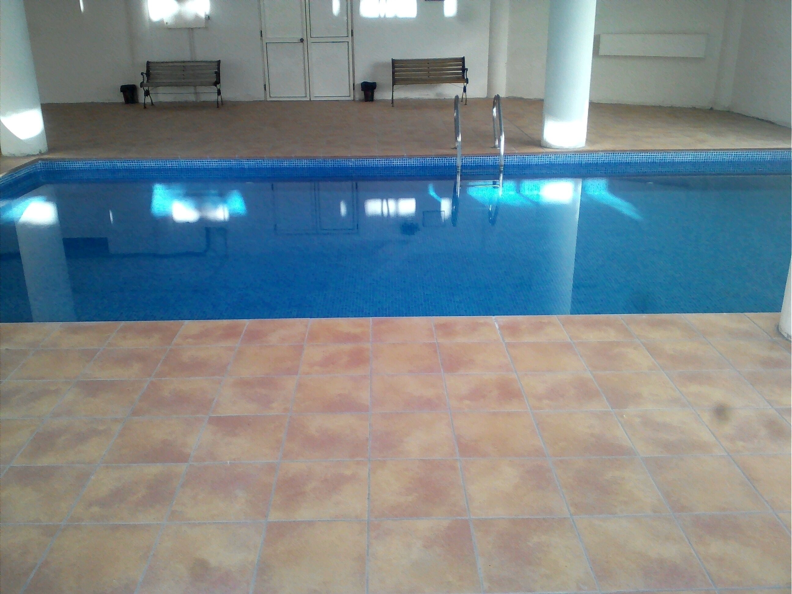 An indoor swimming pool with tiled flooring and benches in the background.