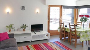 A modern living room with a grey sofa, a flat-screen TV on a white entertainment unit, a colorful striped rug, and a dining area with a green tablecloth. - Zanfleuron A1 by Interhome (Nendaz)
