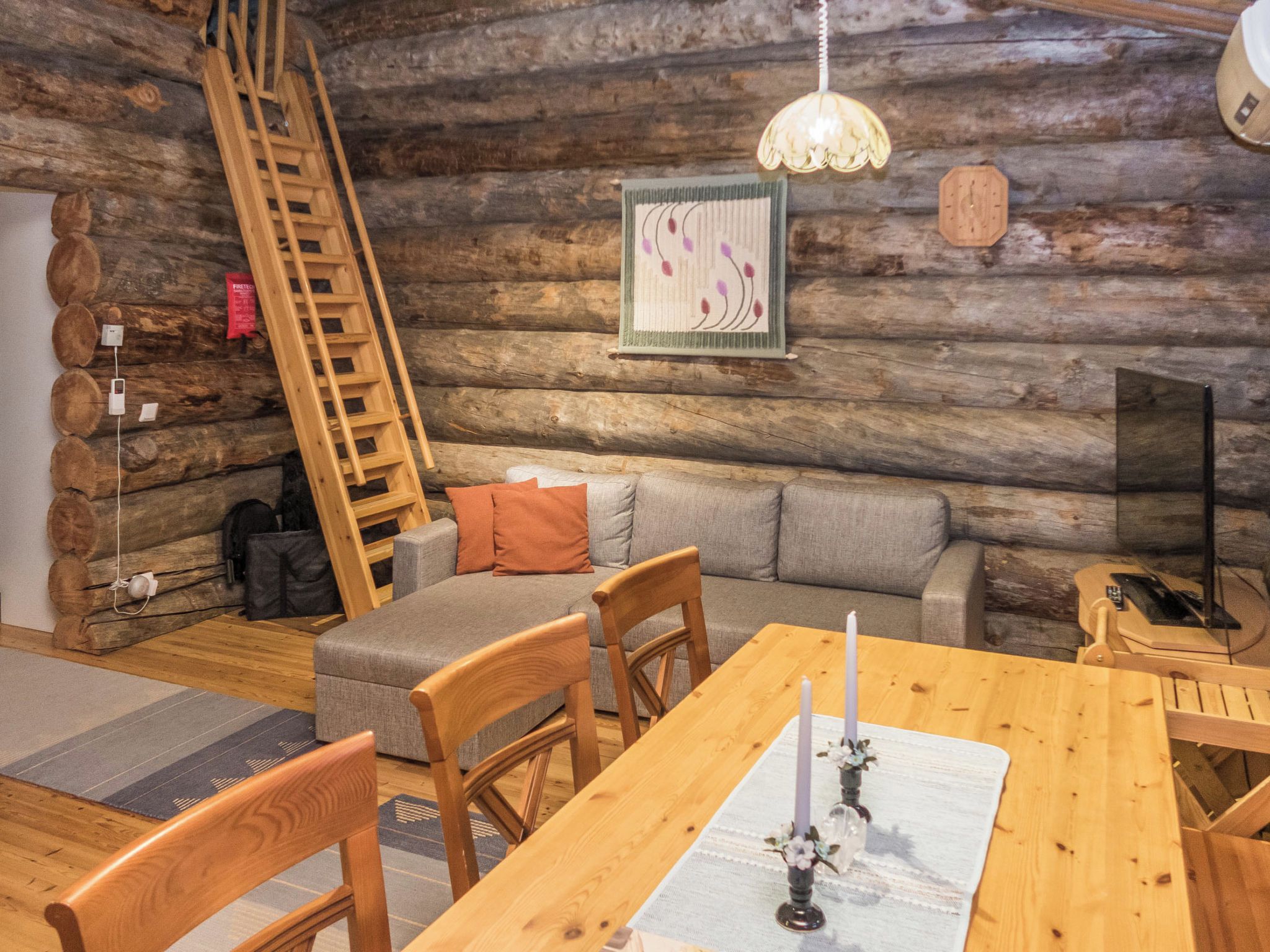 Ein gemütliches Blockhaus-Interieur mit einem Holzesstisch, Stühlen, einer Couch, einer Leiter und einer Hängeleuchte.